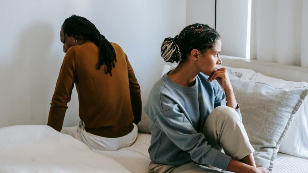 Two people sitting back-to-back on a bed, looking away from each other in a bright room.