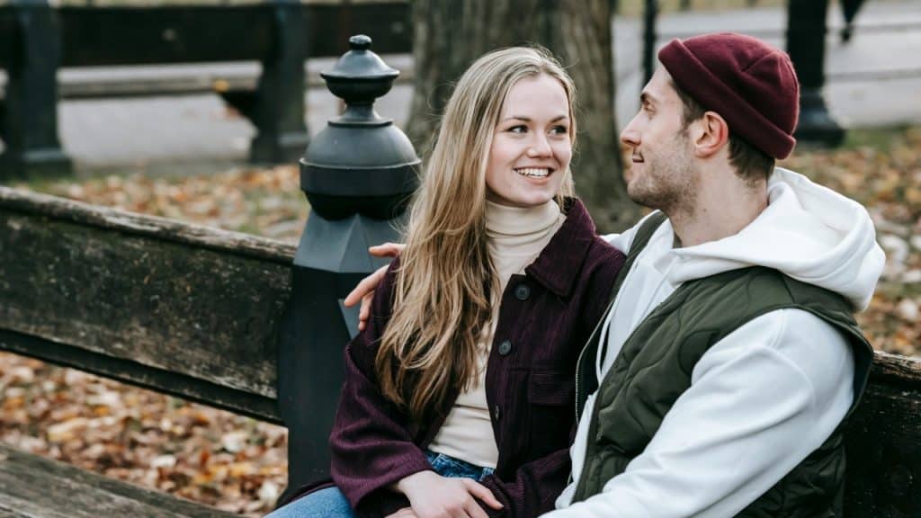 A man and woman sitting on a park bench outdoors, smiling and looking at each other warmly.
