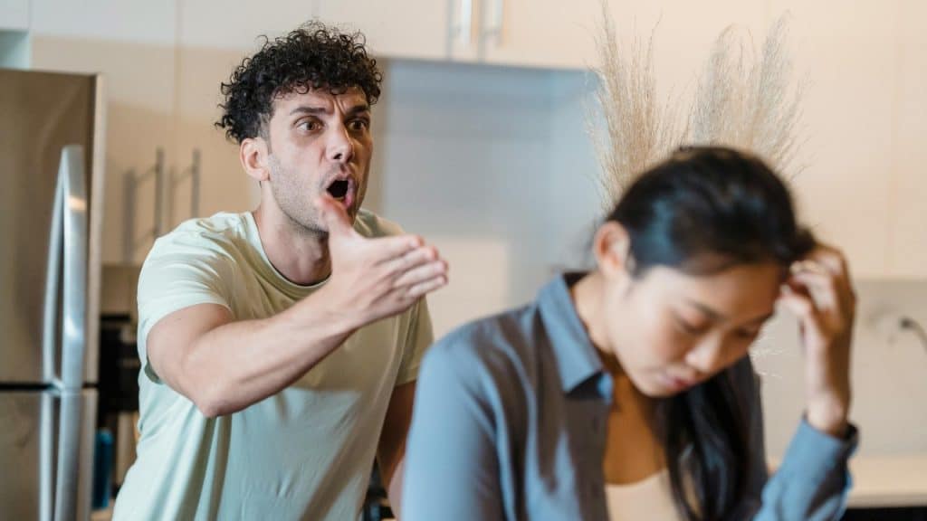 A man gesturing and shouting toward a woman who is looking down and touching her hair.