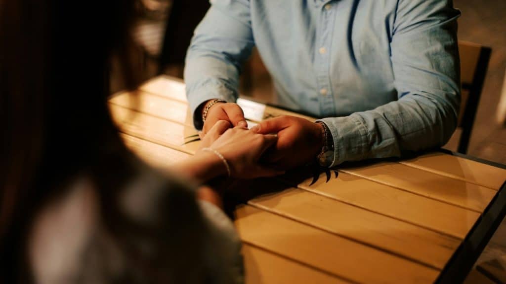 Close-up of two people holding hands across a wooden table in a dimly lit outdoor setting.