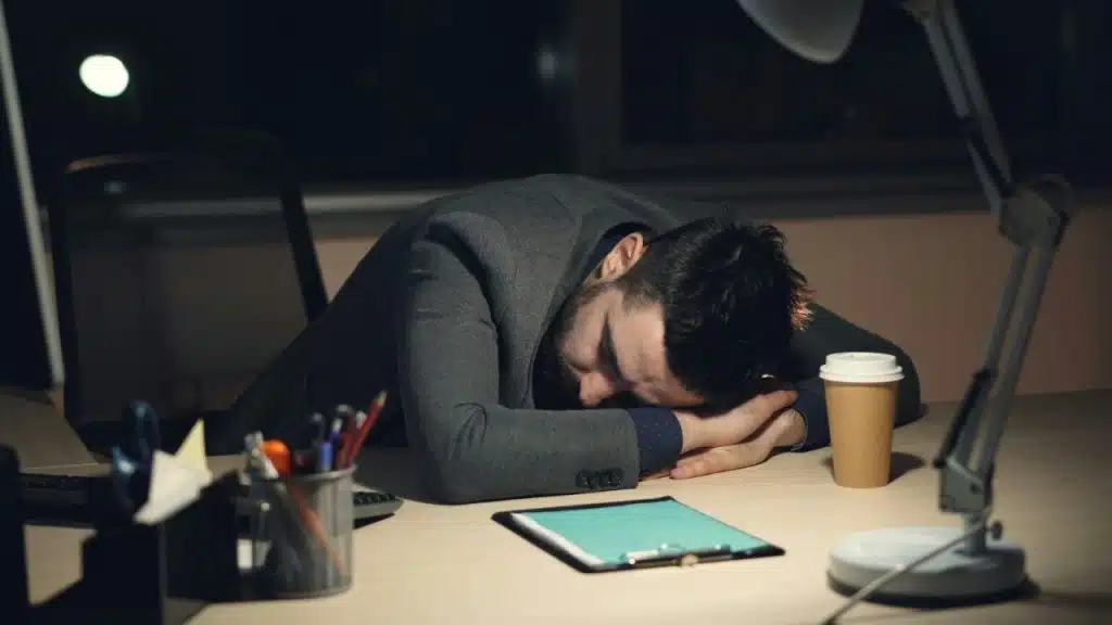 A man in a suit resting his head on his arms on a desk at night.