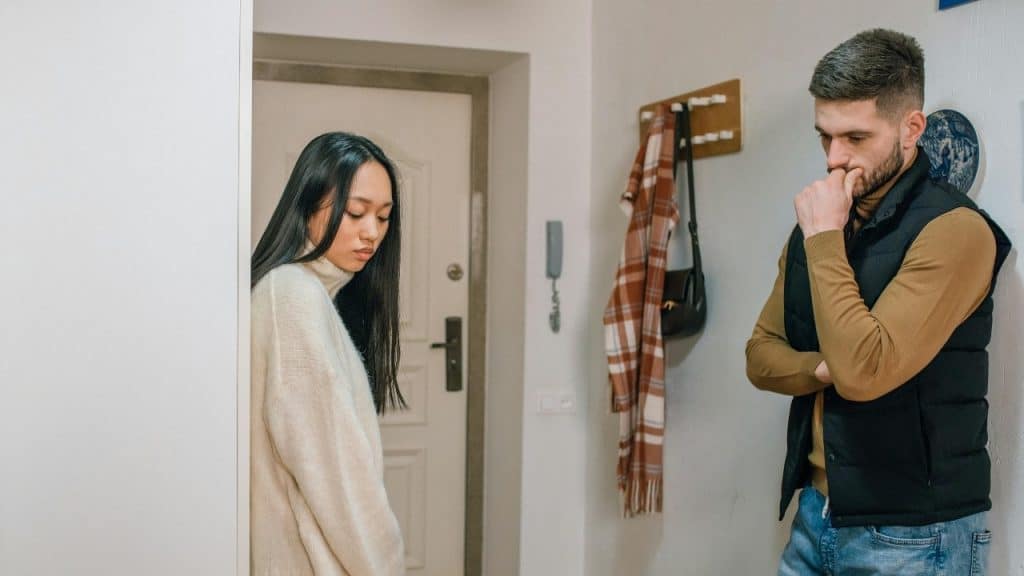 A woman in a white sweater looking down while a man leans against a wall nearby.