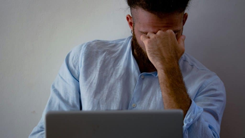 A man with a beard sitting behind a laptop, pressing his hand against his closed eyes.