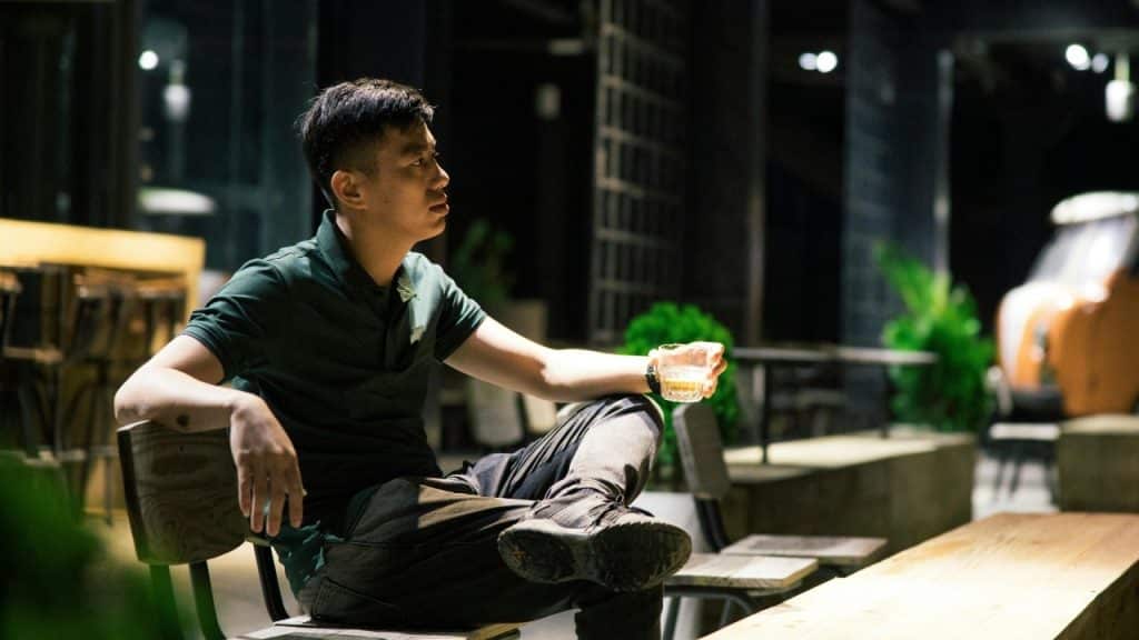 A man sitting outdoors at night, holding a glass and looking off into the distance.
