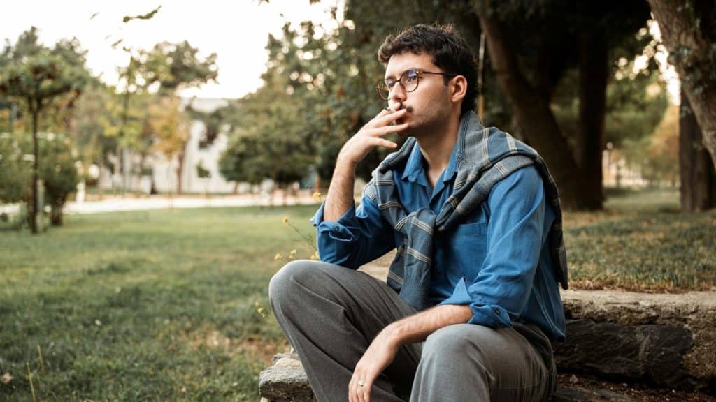 A man with glasses sitting on a stone ledge in a park, holding a lit cigarette.