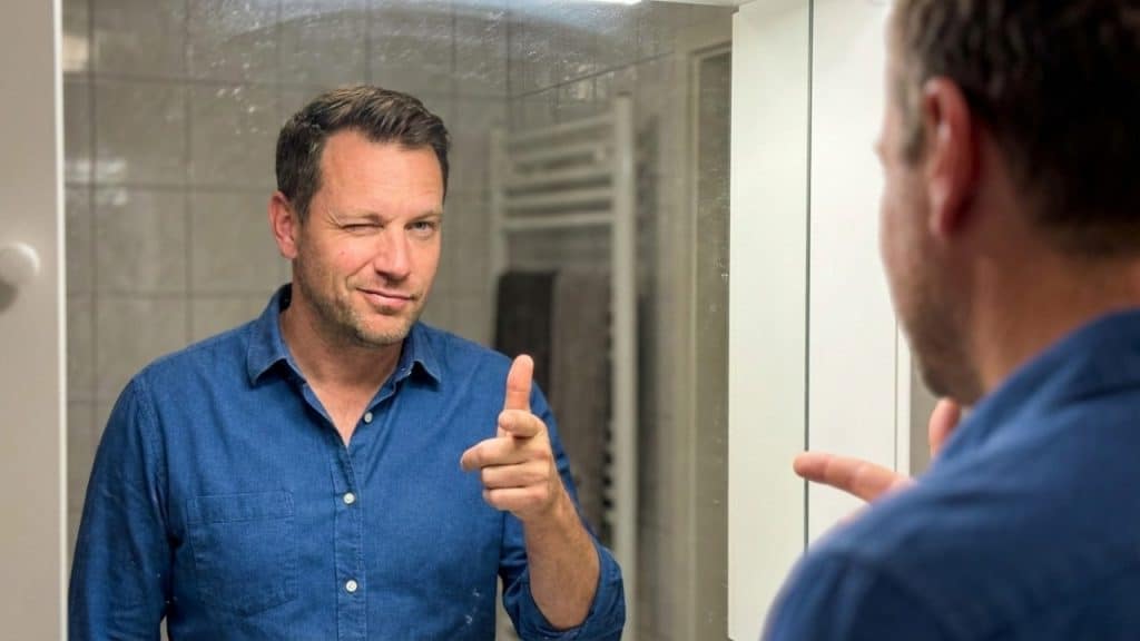 Confident man in a blue shirt winking and pointing at his reflection in the mirror.