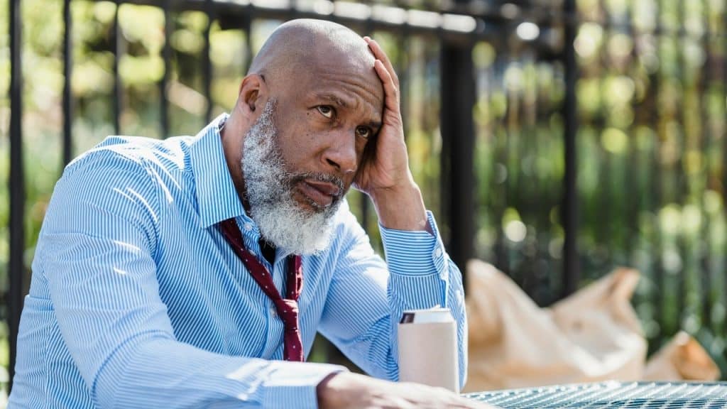 A man with a white beard sits outdoors, resting his head on his hand thoughtfully.