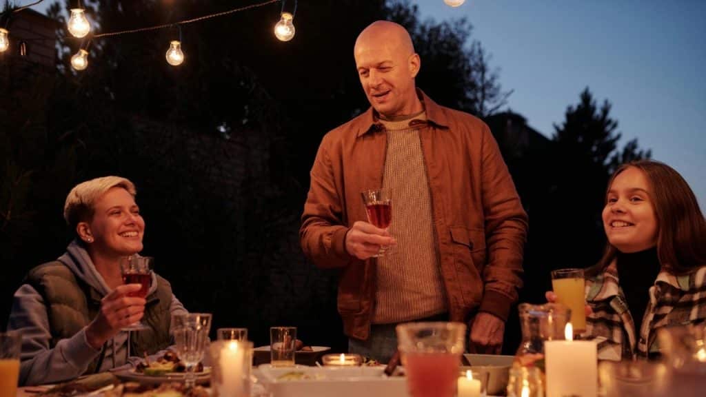 A man stands at a dinner table holding a glass, surrounded by seated, smiling people.
