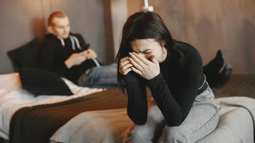 A woman sits on a bed crying into her hands while a man uses a phone.