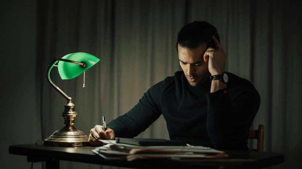 A man sits at a desk under a green lamp, writing on a stack of papers.