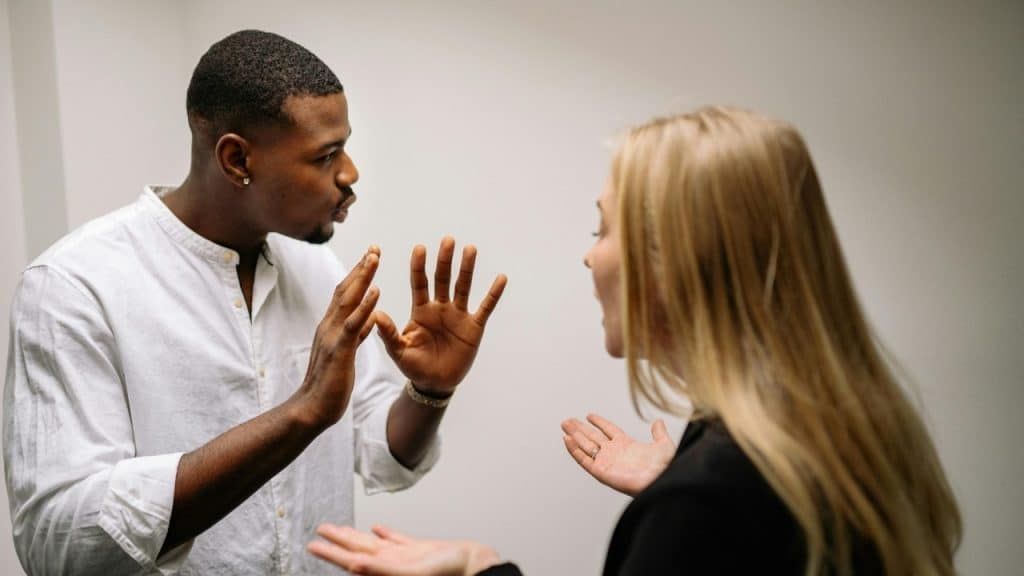 A man in a white shirt raises his hands during a conversation with a blonde woman.