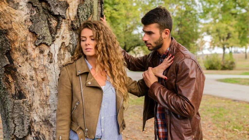 A man leans against a tree near a woman who is pushing his hand away.