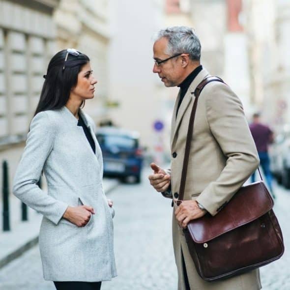 A man and woman in long coats stand on a narrow street talking to each other.