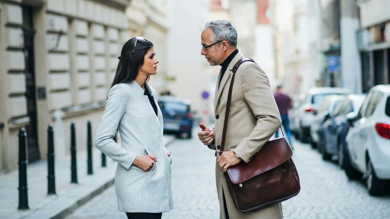 A man and woman in long coats stand on a narrow street talking to each other.