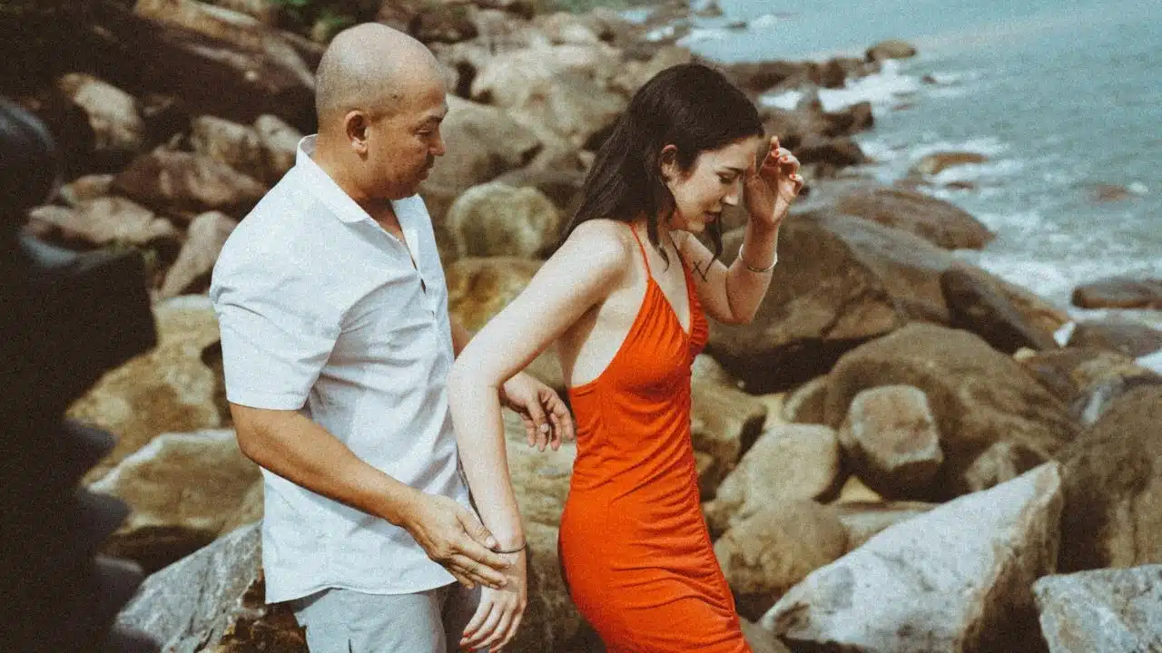 A man in a white shirt assists a woman in a red dress on rocky terrain.