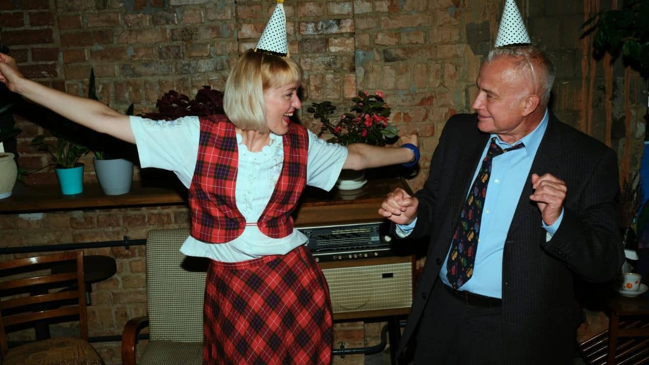 A woman in plaid and a man in a suit dance together wearing polka-dot hats.