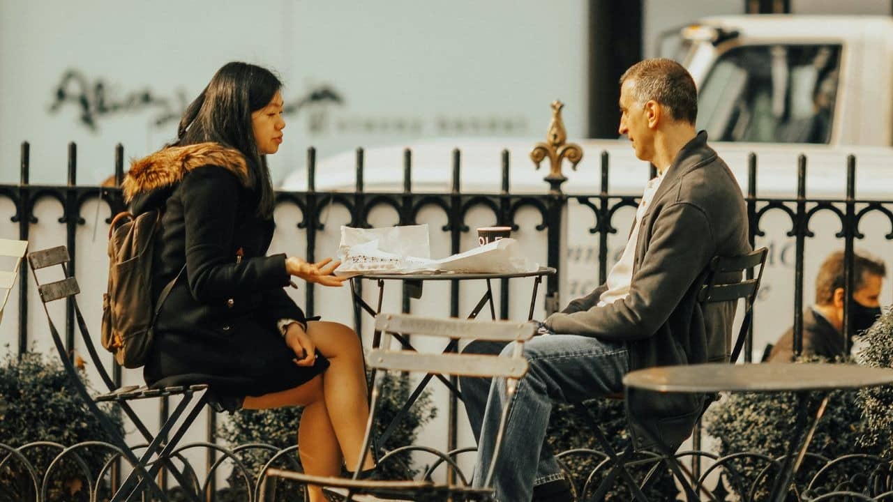 A woman and a man sit at a small outdoor table talking beside a metal fence.