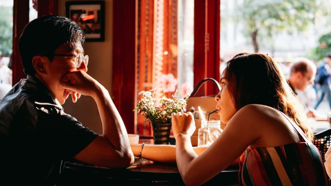 A man and woman sit at a cafe table with flowers between them in sunlight.