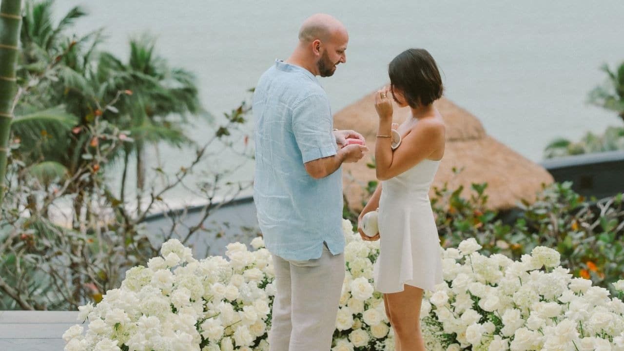 A man holds a small pink box while standing with a woman behind white flowers.