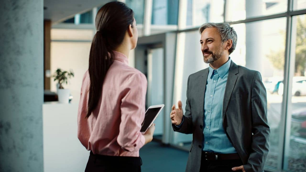 A man in a blazer talks to a woman holding a tablet in a bright office.