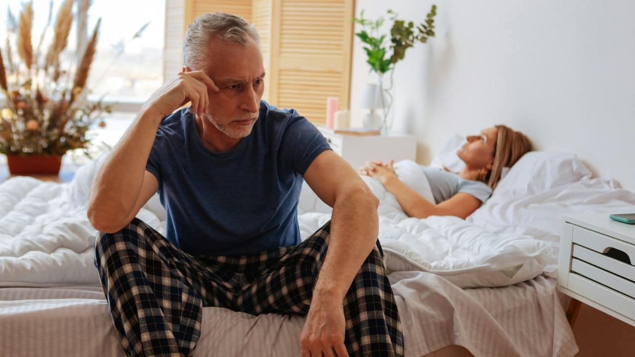 A man sits on a bed looking downcast while a woman lies in the background.