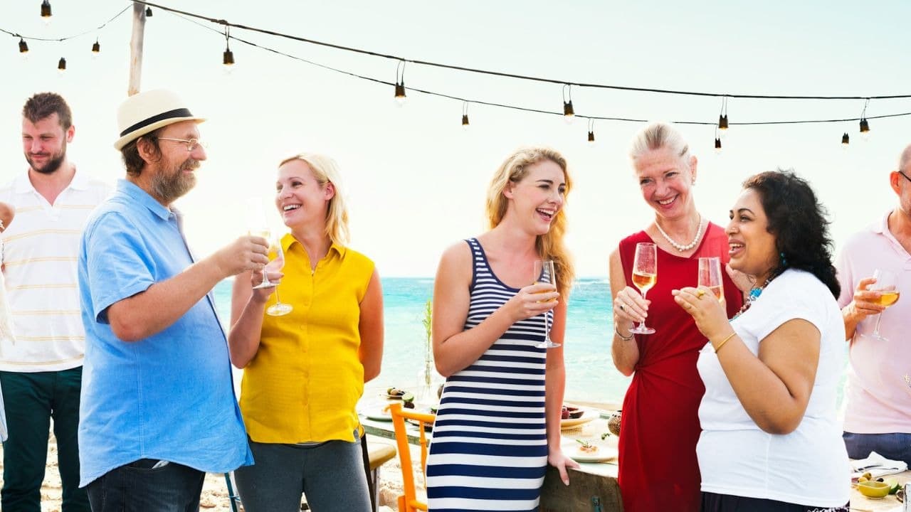 A group of people stand outdoors near the ocean holding glasses and talking near lights.