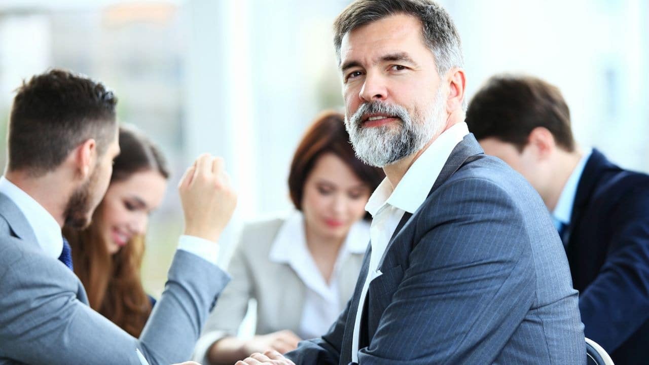 A bearded man in a suit looks forward while others sit at a table behind.