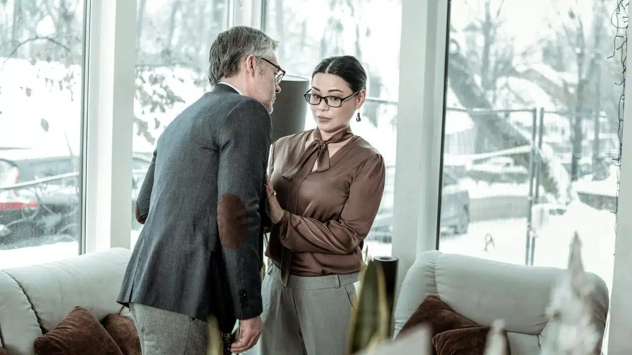 A woman in a brown blouse places her hand on a man's chest near windows.