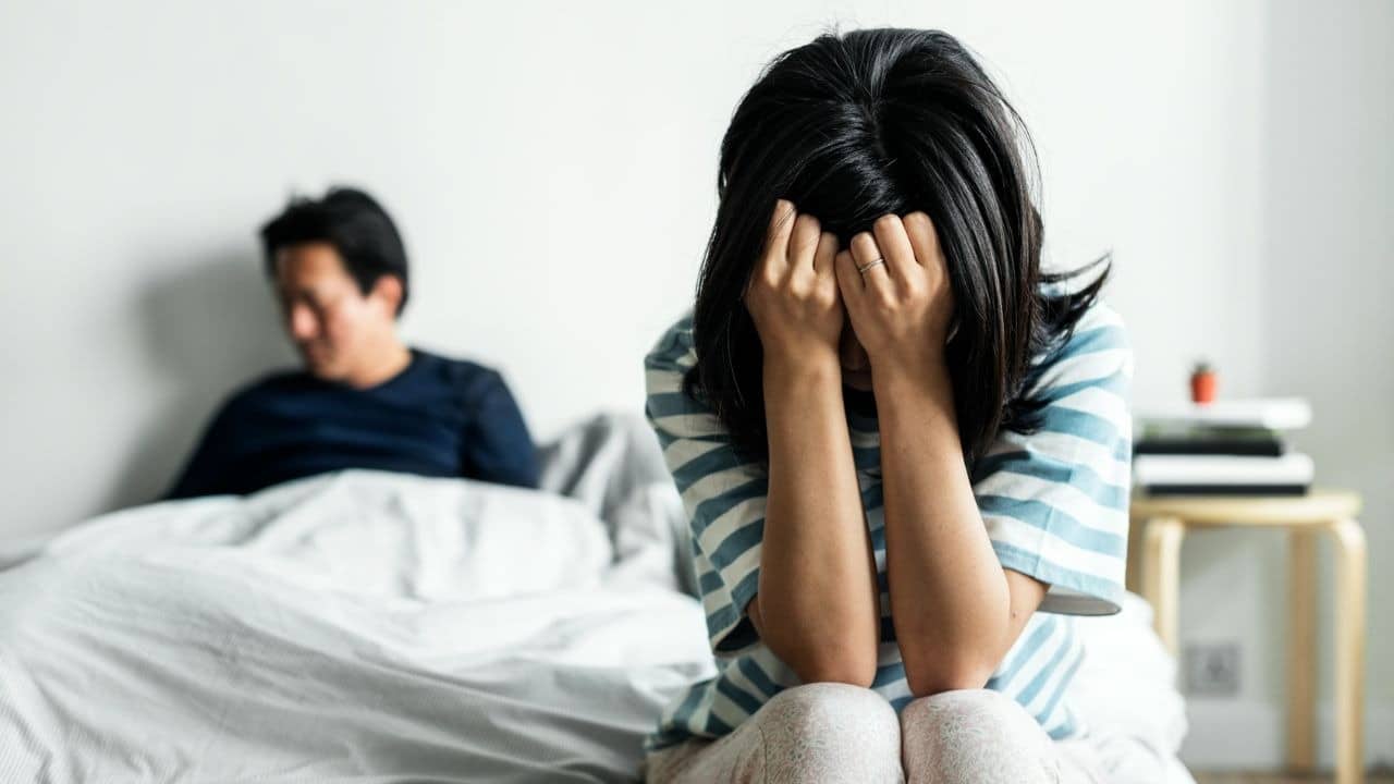 A woman sits on a bed with her head in her hands while a man watches.