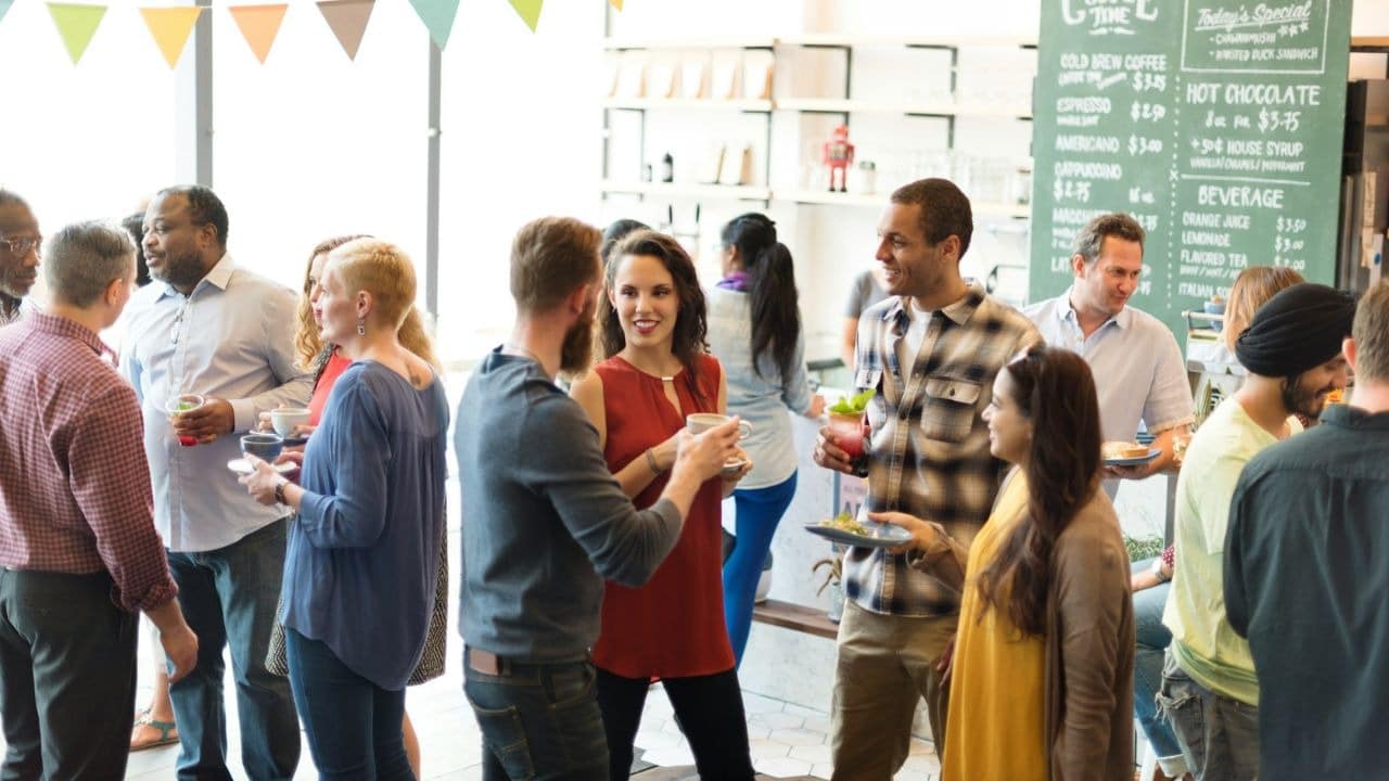 A diverse group of people socialize in a brightly lit cafe with drinks and food.
