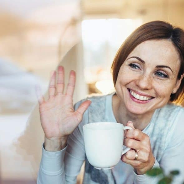 A smiling woman with red hair waves her hand while holding a white ceramic mug.