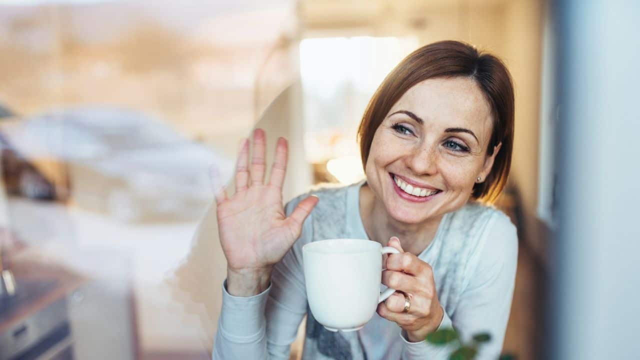 A smiling woman with red hair waves her hand while holding a white ceramic mug.