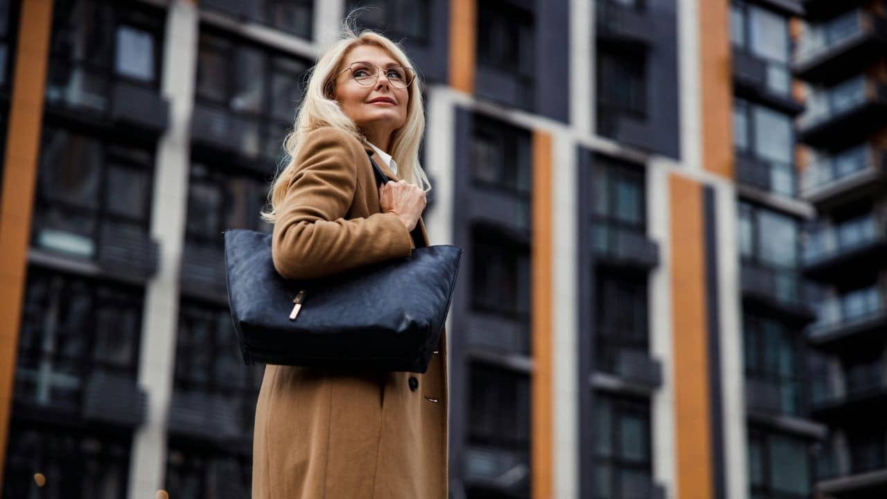 A blonde woman in a tan coat and glasses looks upward before tall urban buildings.