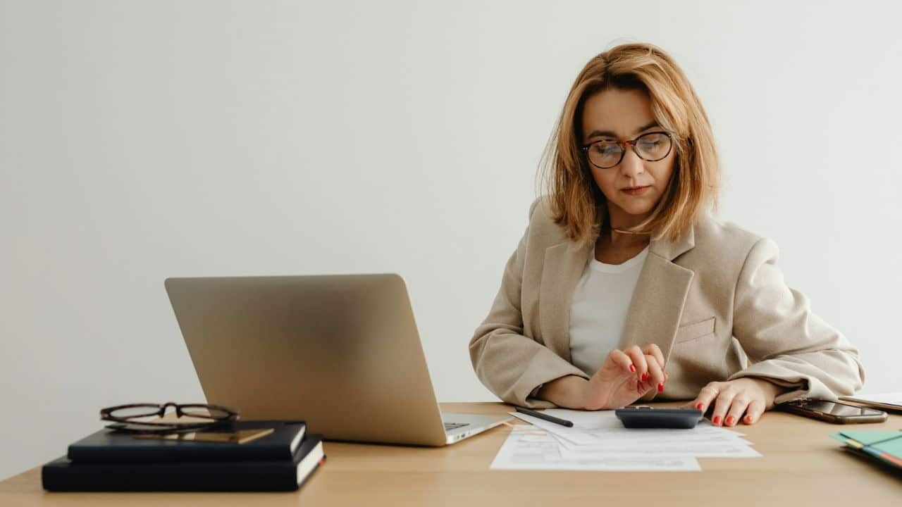 A woman wearing a tan blazer and glasses uses a calculator at a wooden desk.