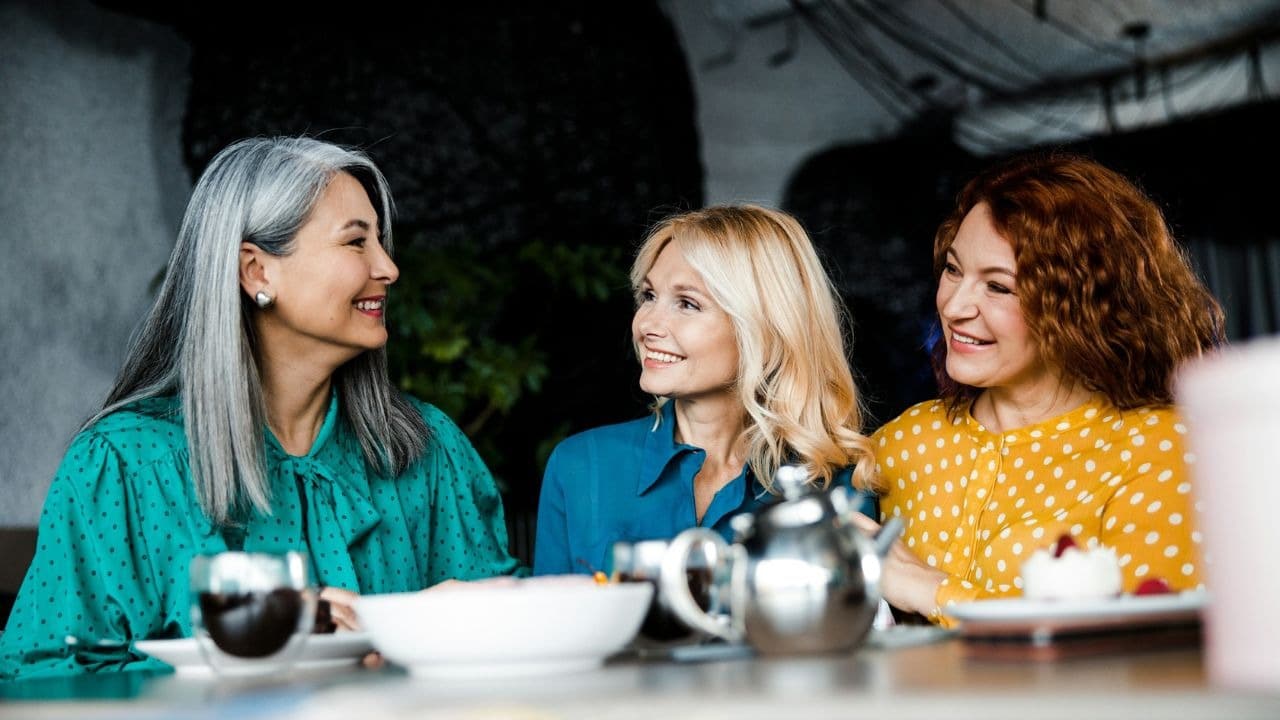 Three women sit at a table together with tea, cake, and a silver metal teapot.