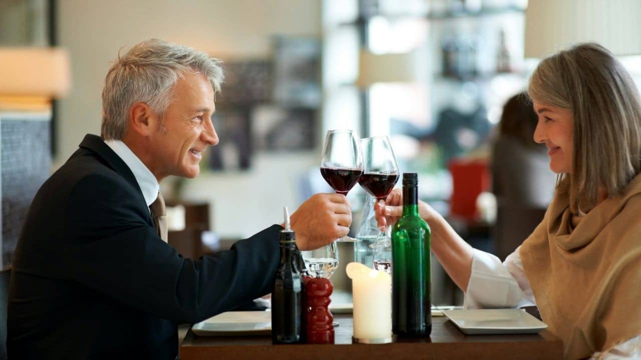A man and woman in formal attire clink wine glasses across a restaurant dinner table.