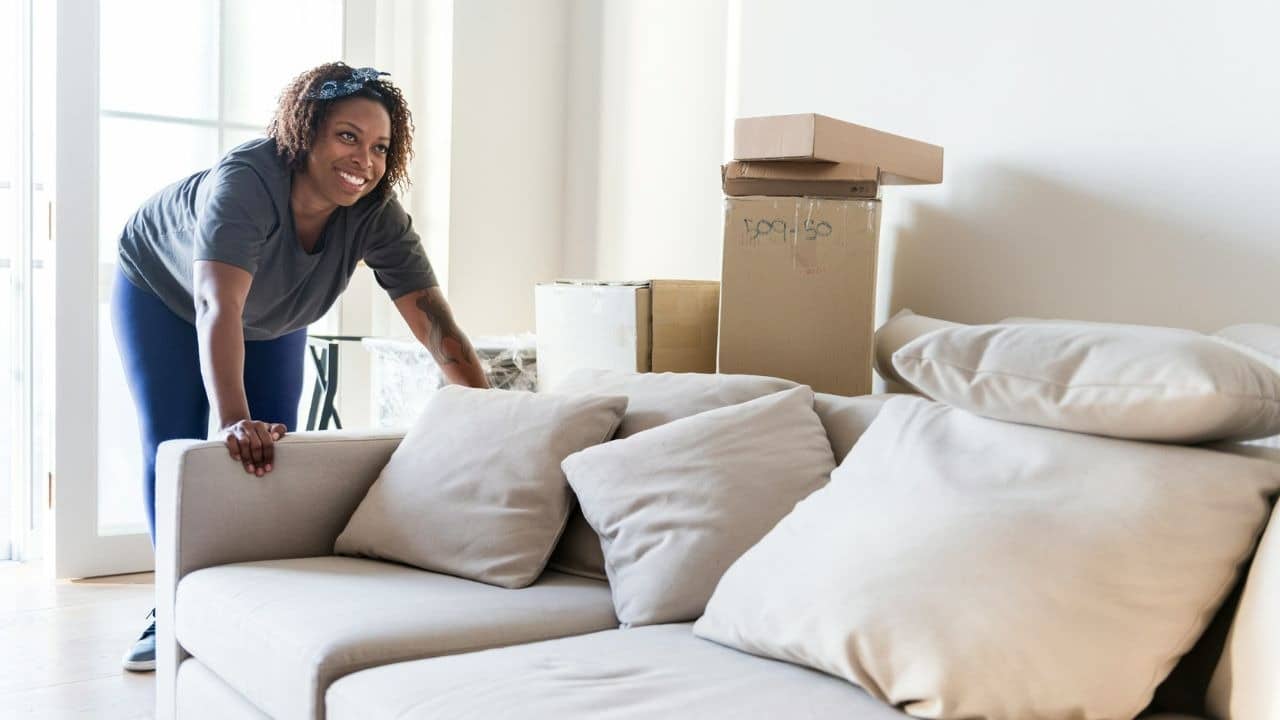 A smiling woman in a headband leans against a sofa near several stacked cardboard boxes.