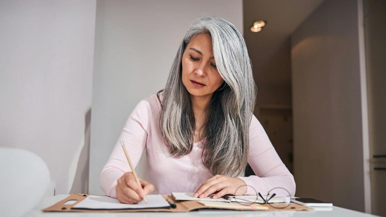 A woman with long gray hair writes in a notebook at a white tabletop surface.