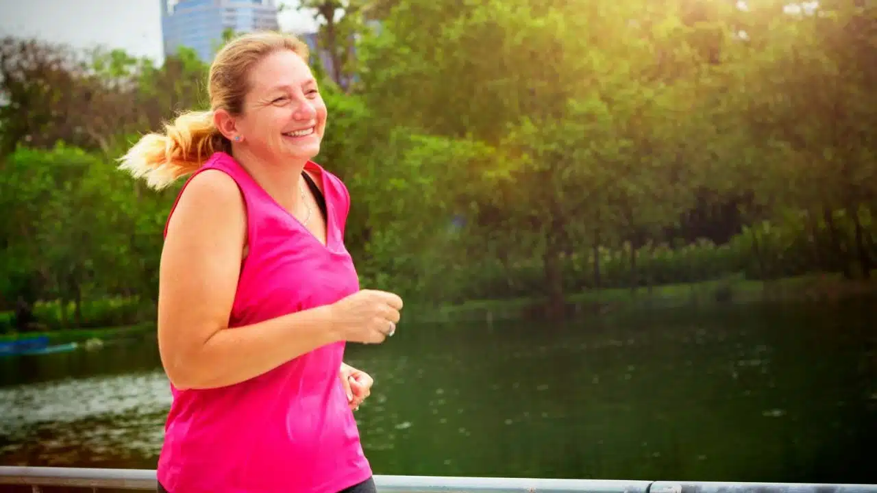 A smiling woman in a pink tank top runs outdoors near a body of water.
