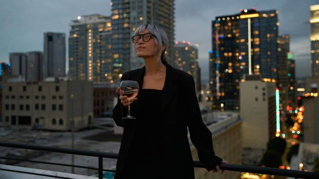 A woman in black clothing and glasses holds a wine glass on an urban rooftop.
