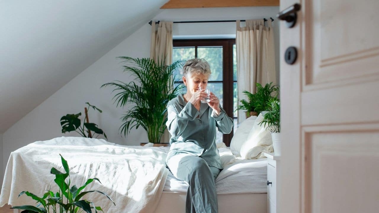 An older woman in green pajamas sits on a bed and drinks from a glass.