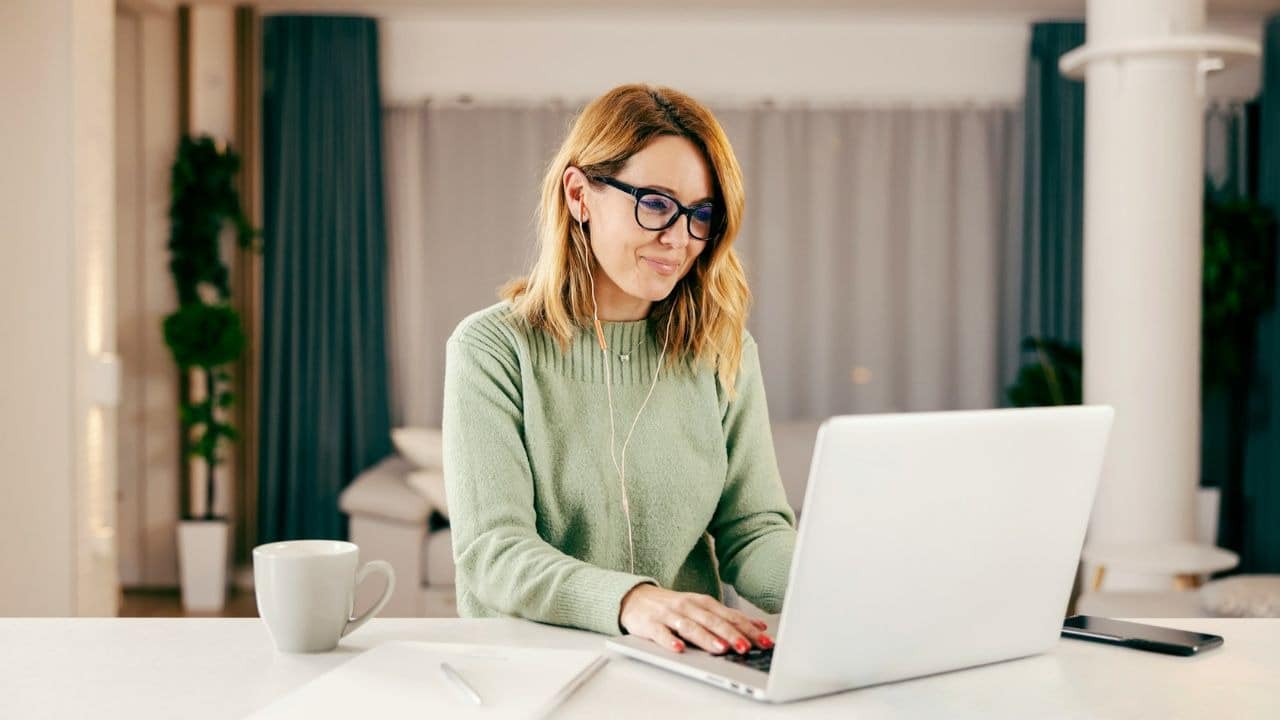 A woman wearing a green sweater and glasses types on a laptop at a table.