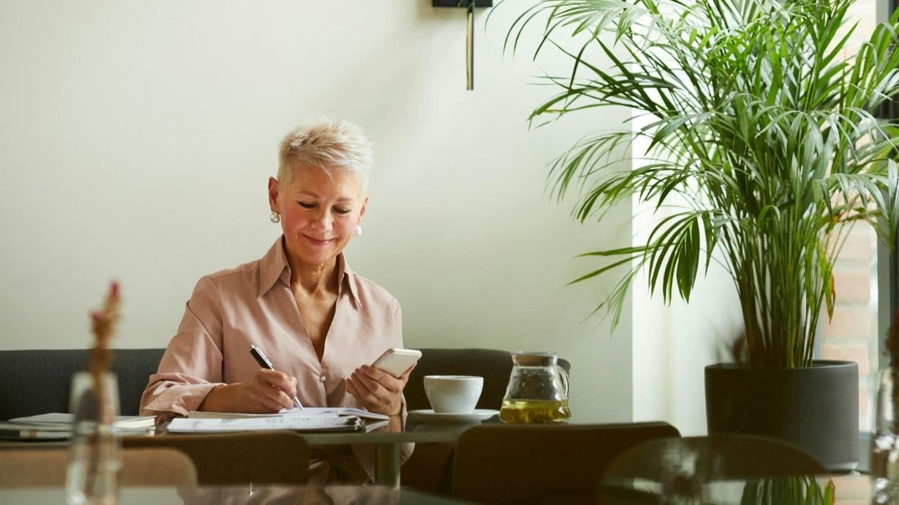 A woman with short blonde hair writes in a notebook while holding a smartphone.