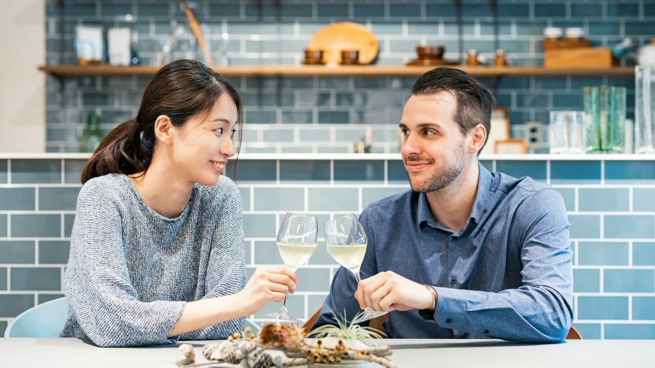 A man and woman clink wine glasses while sitting before a blue tiled wall.