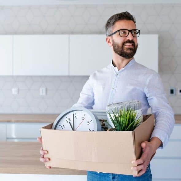 A smiling bearded man in a kitchen holds a cardboard box containing a clock and plant.