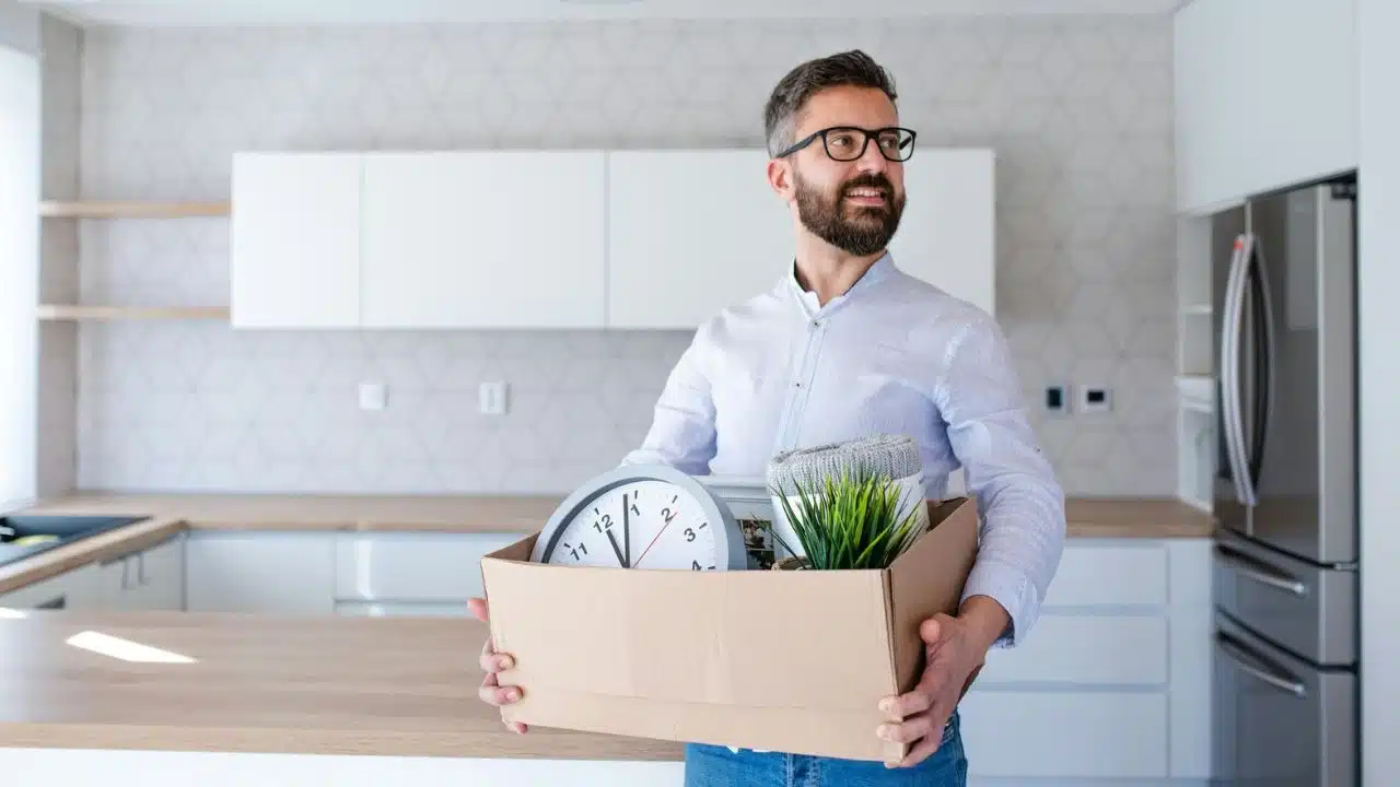 A smiling bearded man in a kitchen holds a cardboard box containing a clock and plant.