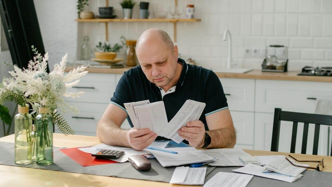 A man sits at a kitchen table reviewing several paper documents with a calculator nearby.