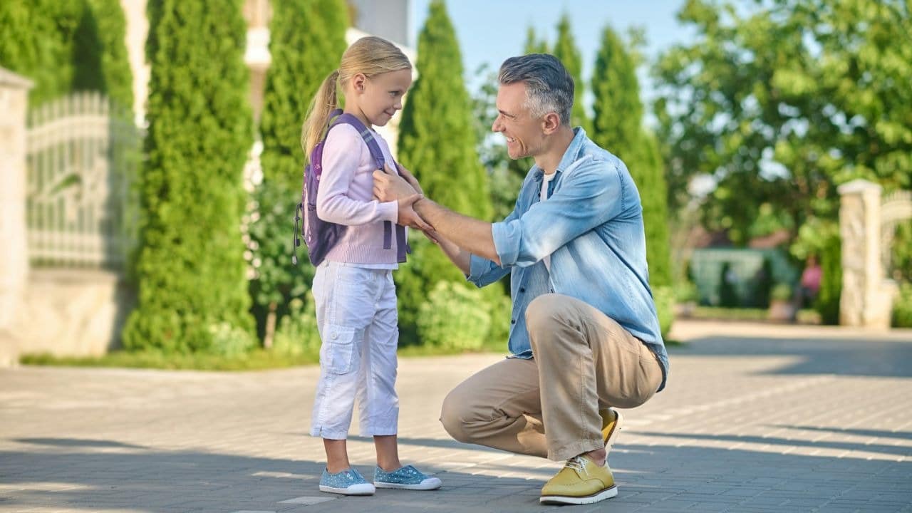 A man kneels on a sidewalk and adjusts the backpack straps of a young smiling girl.