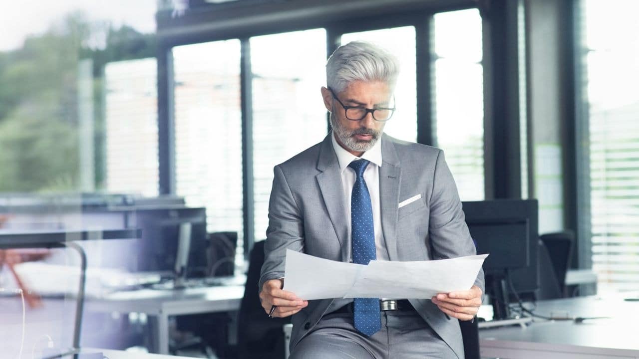 A man with grey hair and glasses wears a suit while reading papers in an office.