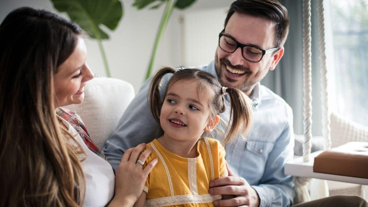 A man and woman smile while sitting on a couch with a young girl in pigtails.